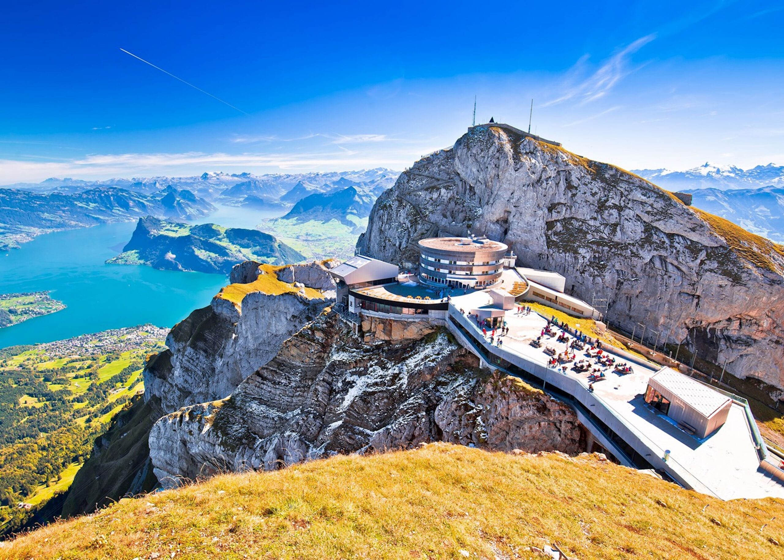 Circular, modern building on a clifftop with people on a viewing terrace against a blue alpine sky.