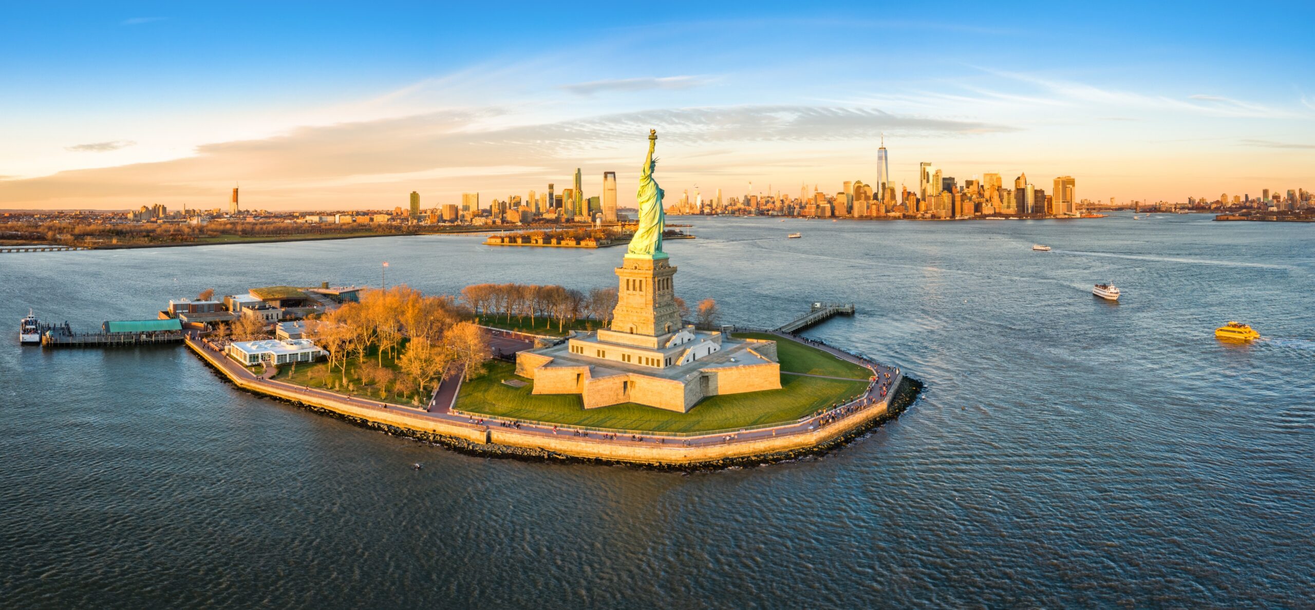 Aerial view of the Statue of Liberty on Liberty Island with the New York City skyline across the harbor at sunset/sunlit.