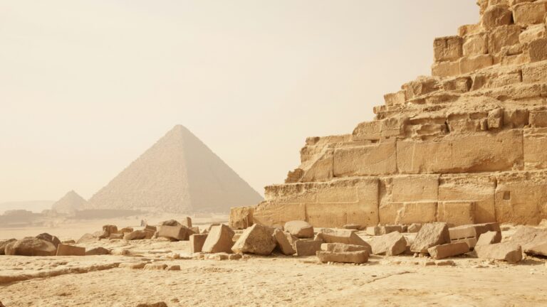 Desert scene with weathered limestone ruins in the foreground and the pyramids in the hazy distance under a warm sky.