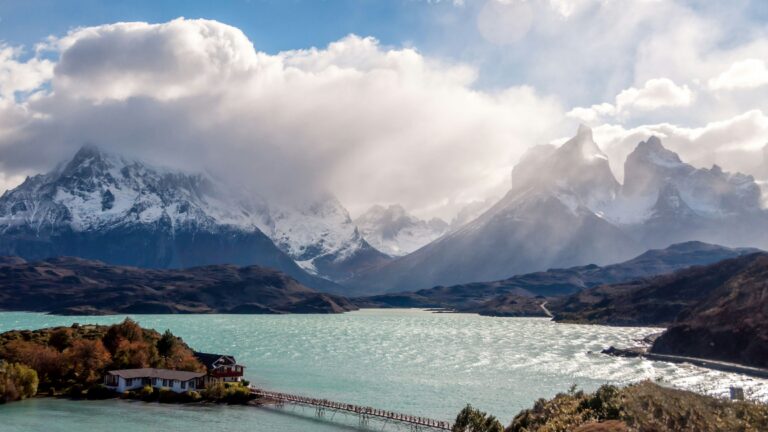 Snowy mountain range behind a turquoise lake with a small lakeside village and a wooden bridge in the foreground.