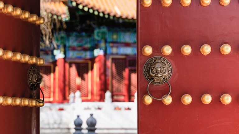 Close-up of a red wooden temple gate with gold studs and a brass lion door knocker, opening to a blurred traditional courtyard.