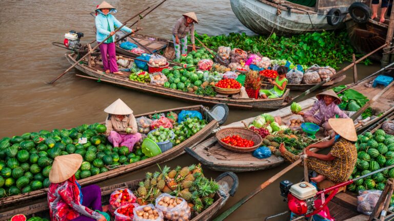 Floating market scene with multiple boats loaded with colorful fruits and vegetables along a brown river, vendors wearing conical hats.