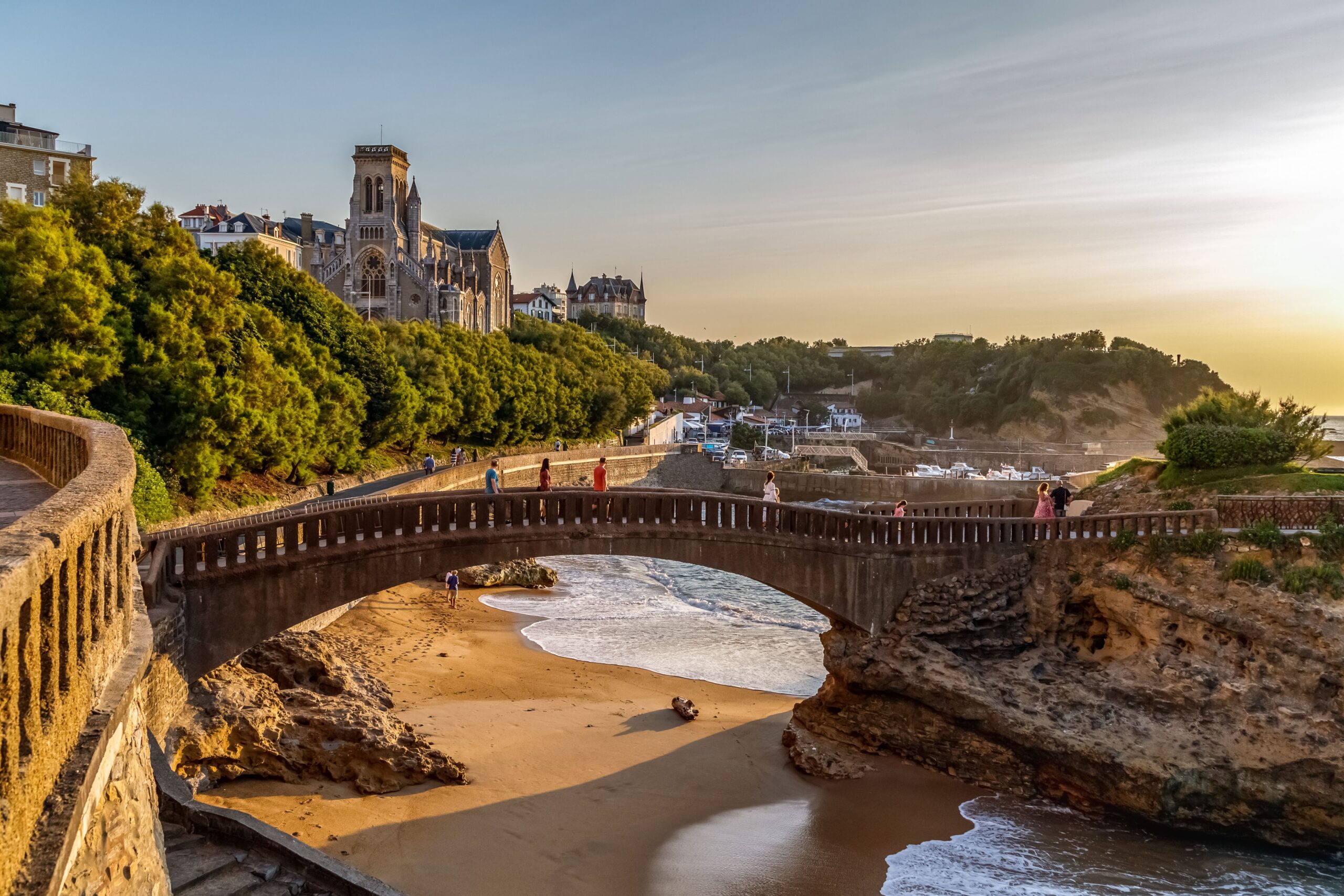 Coastal town with a Gothic church perched on a hillside, a stone curved bridge over a sandy cove at sunset.