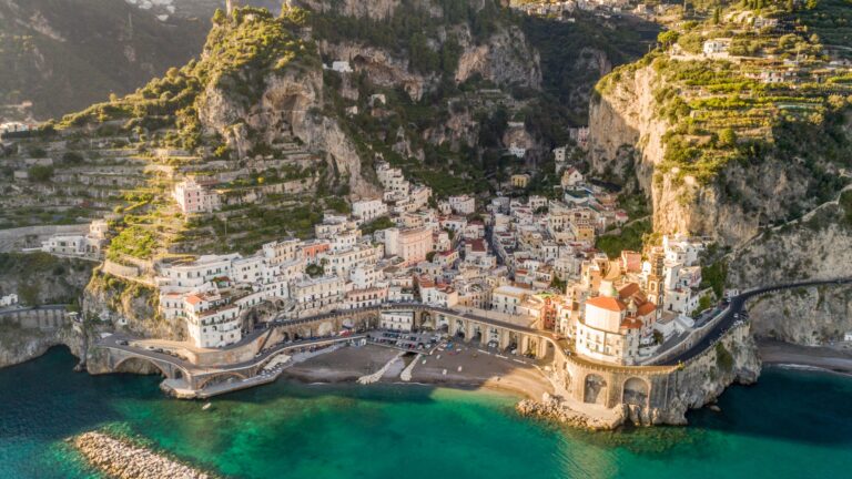 Aerial view of a cliffside Italian village with pastel houses built into the rocky coast and a curved seawall along turquoise water at the shore.