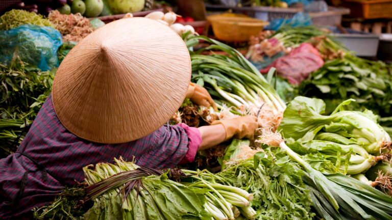 Vendor wearing a conical straw hat arranging fresh leafy greens at a busy market stall.