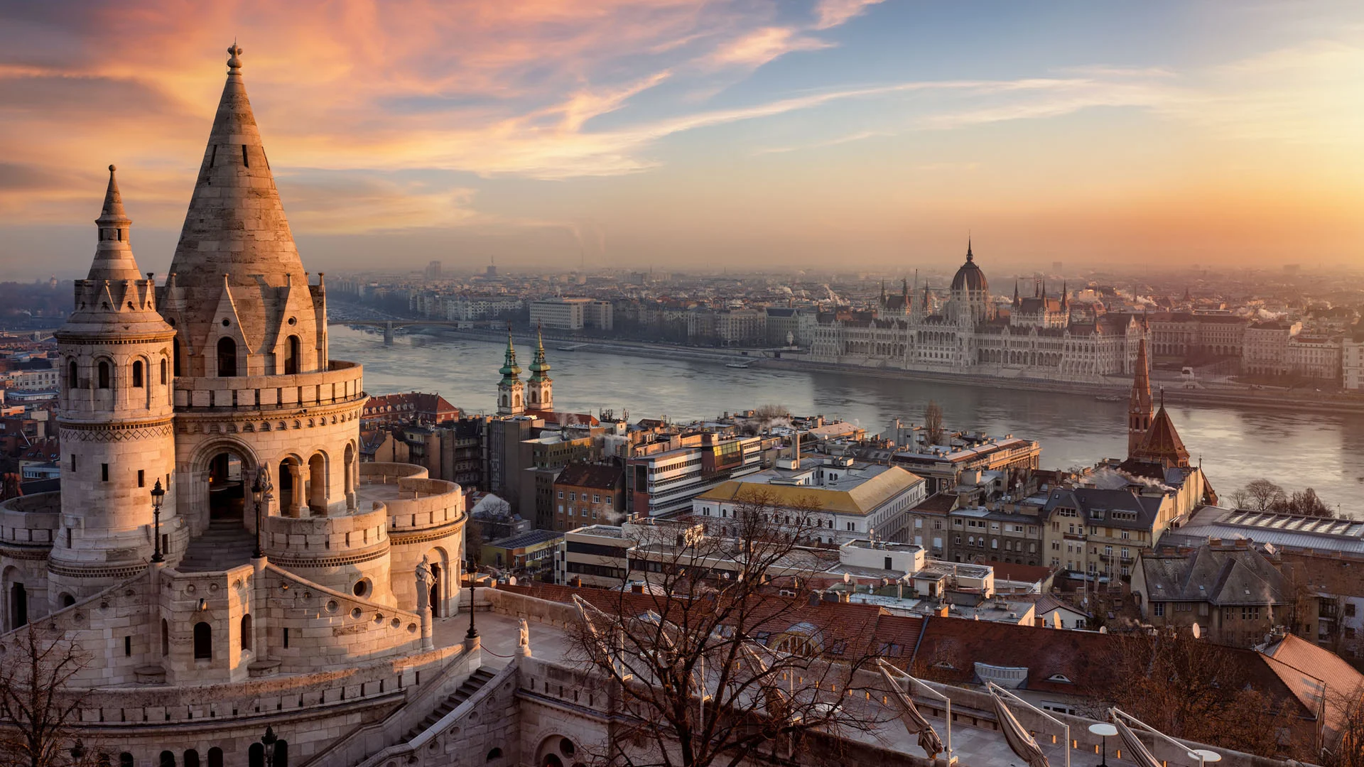 Panoramic view of Budapest at sunset, with the Fisherman’s Bastion towers in the foreground and the Danube River stretching across the cityscape behind it.