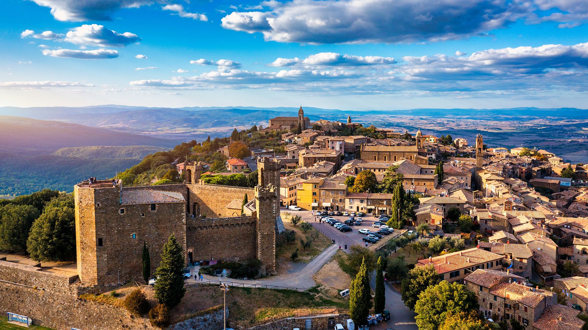 Panoramic view of a medieval hill town with stone buildings and towers atop a hillside, under a blue sky with scattered clouds.