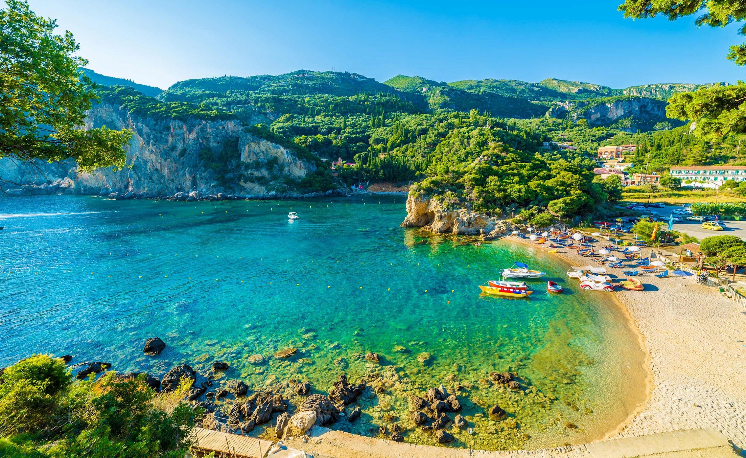 Sunny coastal cove with clear turquoise water, rocky headlands, and a pebbled beach lined with umbrellas and small boats.