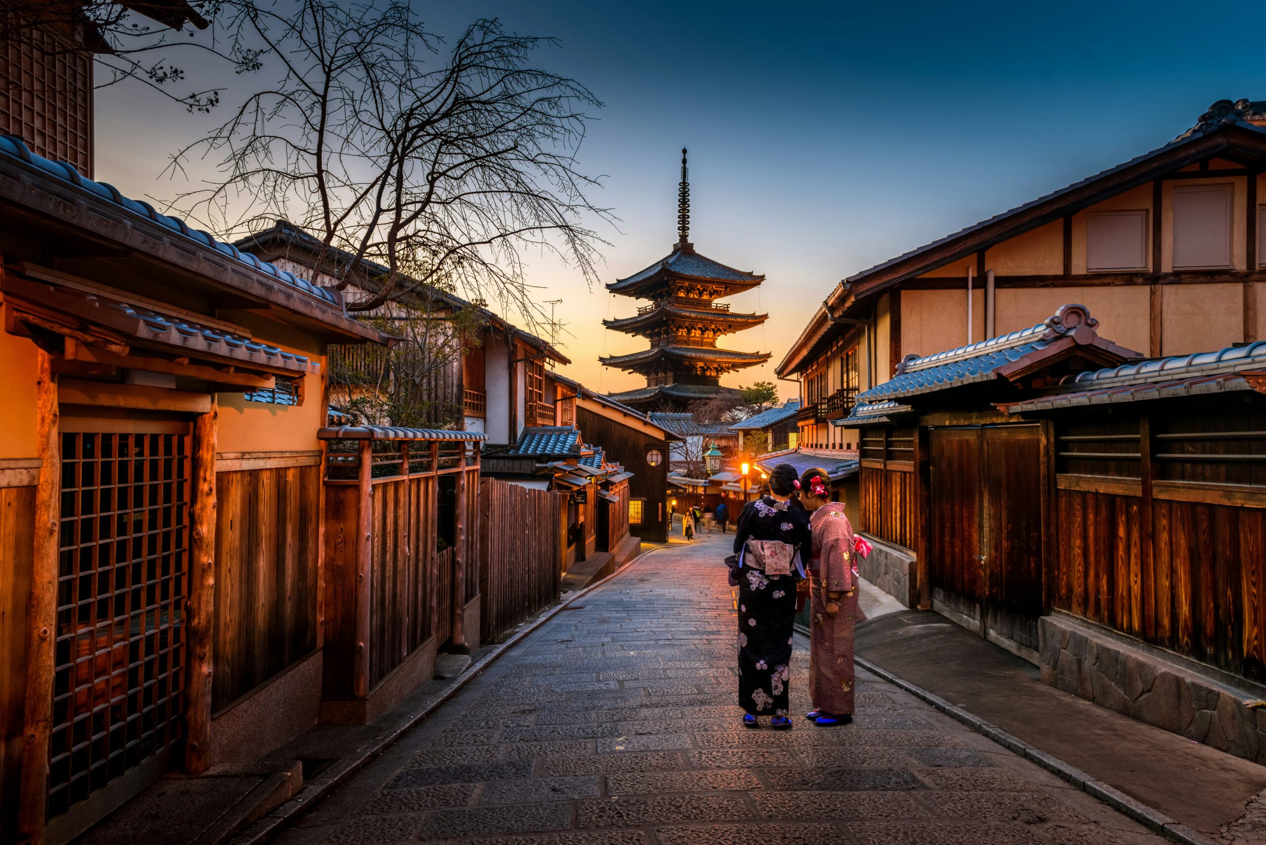 Traditional wooden street with a three-tier pagoda in the distance at sunset; two women in kimonos walk together in foreground.